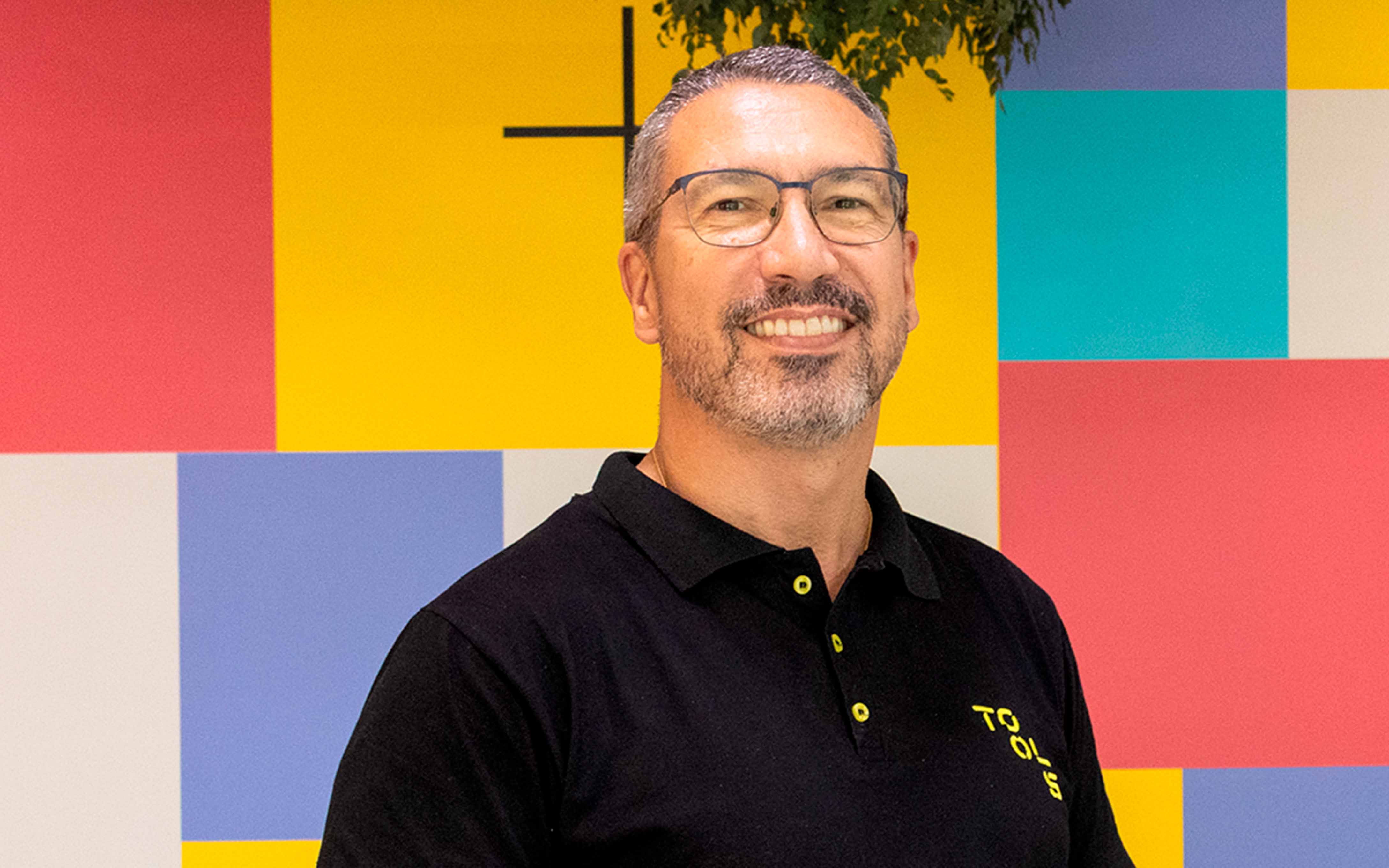 Homem branco, grisalho, de óculos, sorrindo utilizando a camiseta polo preta da TOOLS Digital Services.