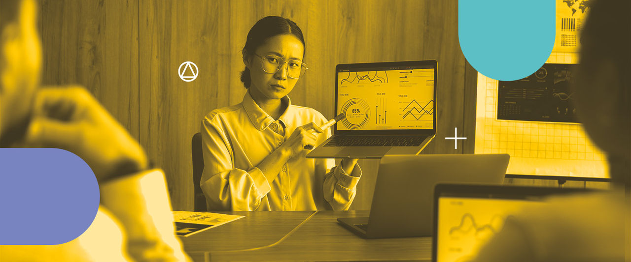 Mulher asiática, de óculos e cabelos presos, segura um notebook e aponta para o gráfico da tela com uma caneta.